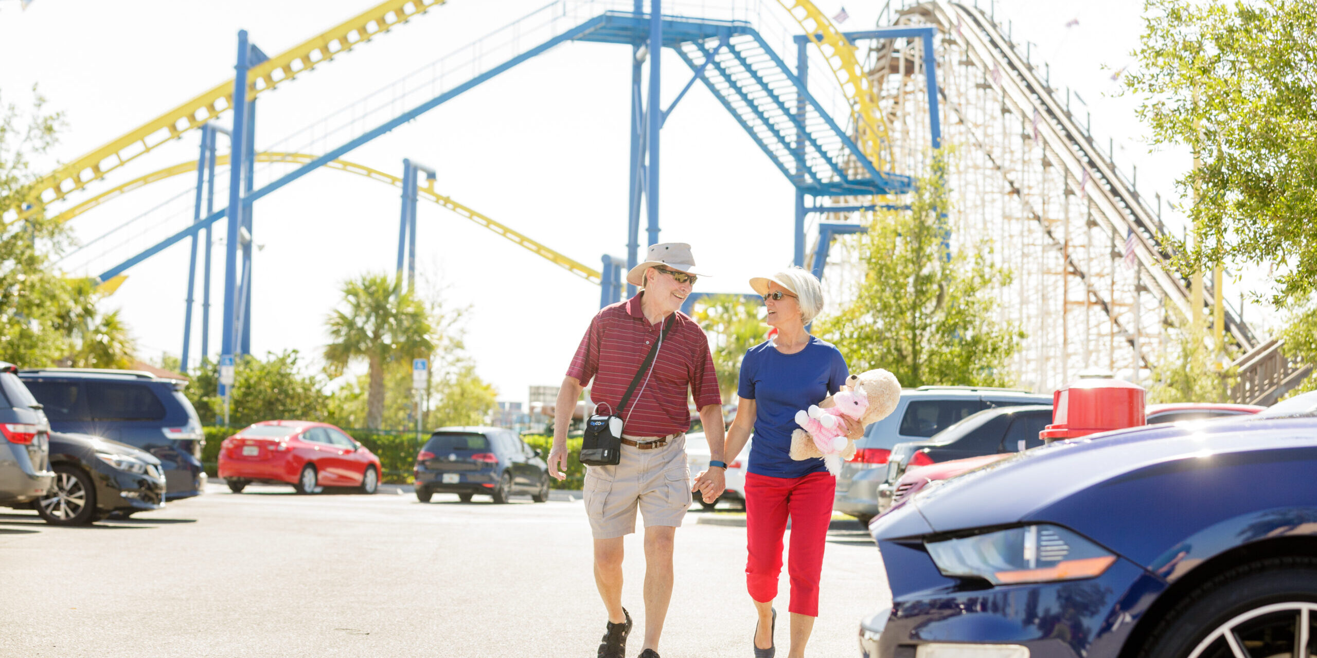 Photo of a couple at an amusement park. The husband is carying a small portable oxygen concentrator and the wife is carrying two stuffed animal toys.